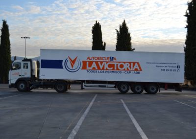 Imagen del perfil de un camión blanco de la autoescuela la victoria enganchado a un tráiler de carga