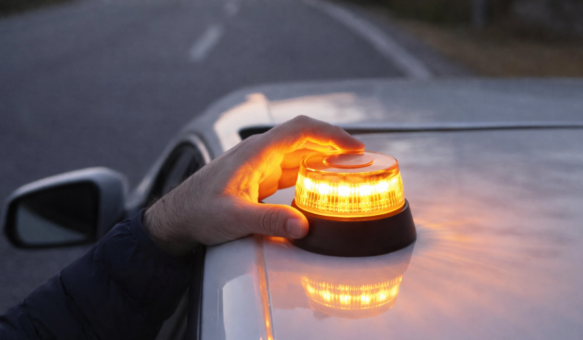 Mano colocando una baliza V16 encendida sobre el techo de un coche blanco en una carretera